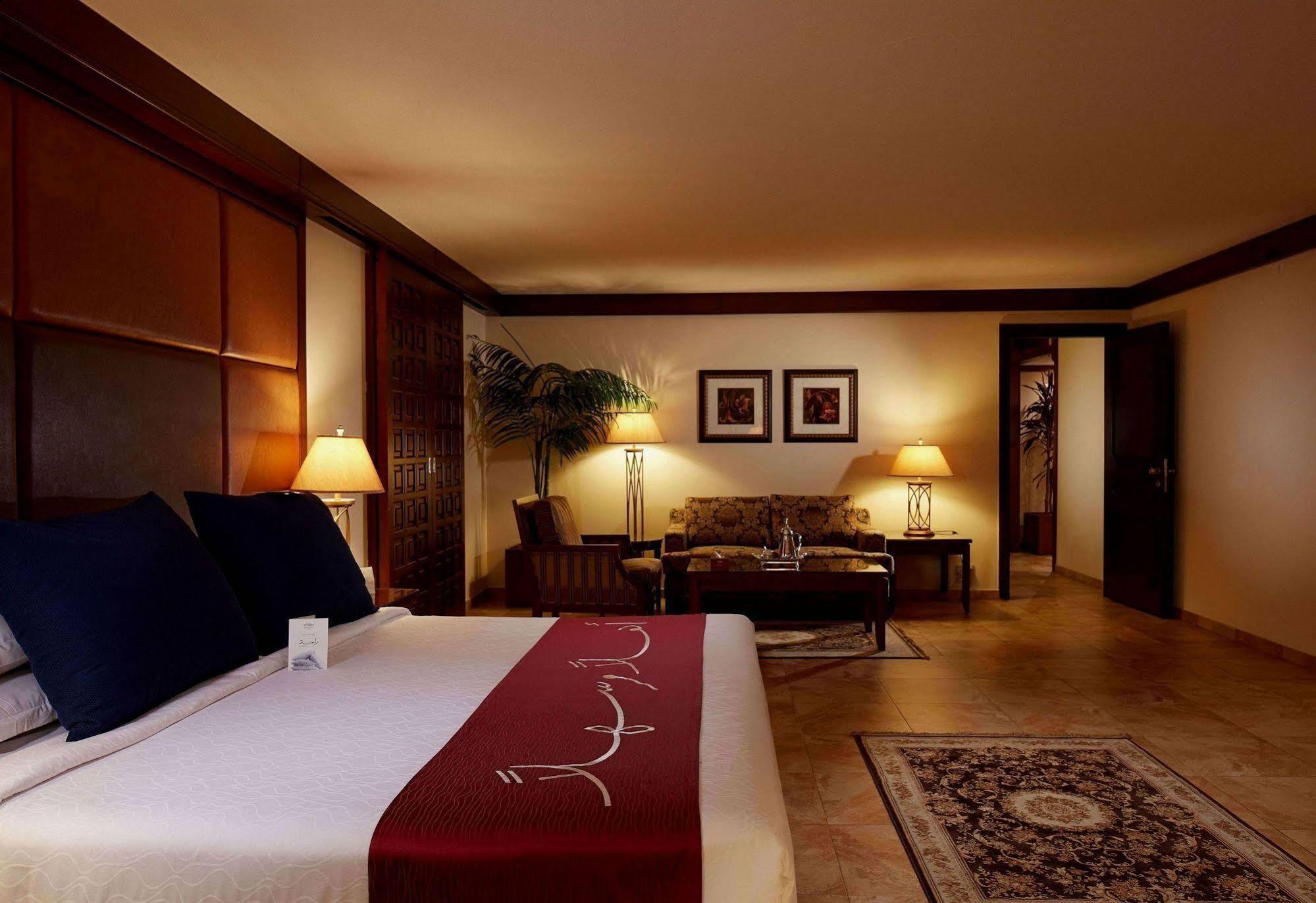 מכה Makarem Ajyad Makkah Hotel מראה חיצוני תמונה The photo depicts a cozy hotel room with a warm and inviting atmosphere. It features a large bed with dark blue pillows and a maroon throw blanket. On either side of the bed, there are lamps providing soft lighting. In the background, a small seating
