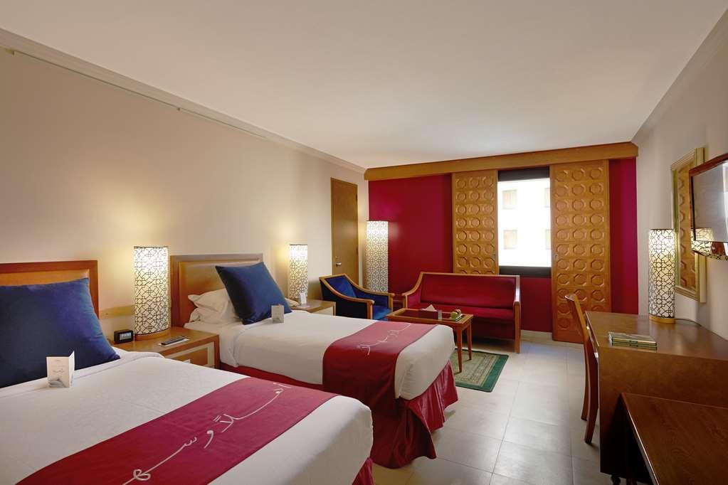 מכה Makarem Ajyad Makkah Hotel חדר תמונה The photo shows a hotel room featuring two twin beds with red and white bedding. There are decorative lamps on either side of the beds. A small seating area is visible, including a blue chair and a red sofa. There is a wooden desk against one wall, a