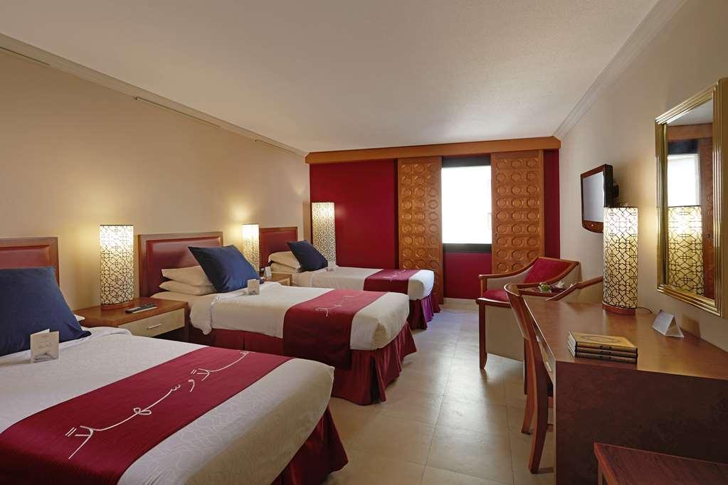 מכה Makarem Ajyad Makkah Hotel חדר תמונה The photo shows a hotel room featuring three beds arranged with red and white bedding. The walls are painted in a neutral tone, and there are decorative lamps on nightstands beside each bed, providing ambient lighting. One of the walls has a window c