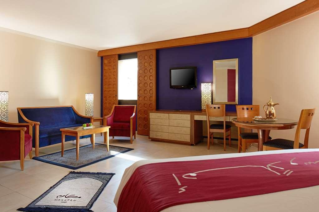 מכה Makarem Ajyad Makkah Hotel חדר תמונה The image shows a hotel room with a modern, comfortable design. The room features a large bed with a red and white bedspread, a seating area that includes a blue sofa, two red chairs, and a wooden coffee table. There's a flat-screen TV mounted on the