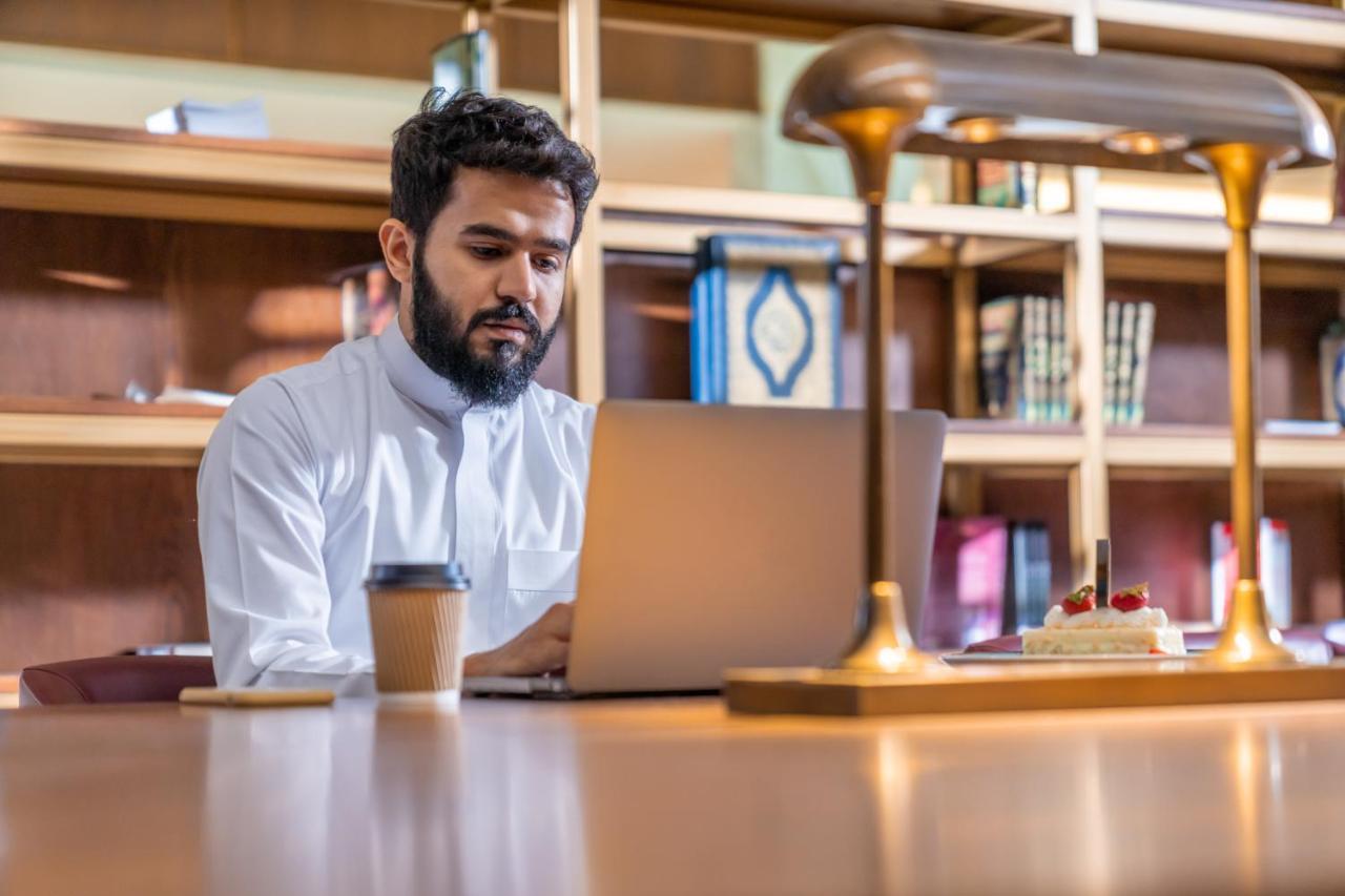 מכה Makarem Ajyad Makkah Hotel מראה חיצוני תמונה The photo shows a man sitting at a desk in a library or a study area. He is wearing a white traditional garment and is focused on working on a laptop. In front of him, there is a cup of coffee and a small cake decorated with strawberries. The backgro