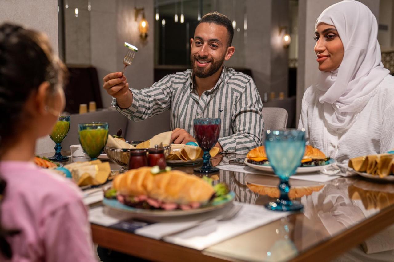 מכה Makarem Ajyad Makkah Hotel מראה חיצוני תמונה The photo shows a dining scene with three people enjoying a meal together. A man is seated at the table, smiling and holding a utensil, possibly ready to serve or share food. He is dressed in a striped shirt. Across from him is a woman wearing a whit