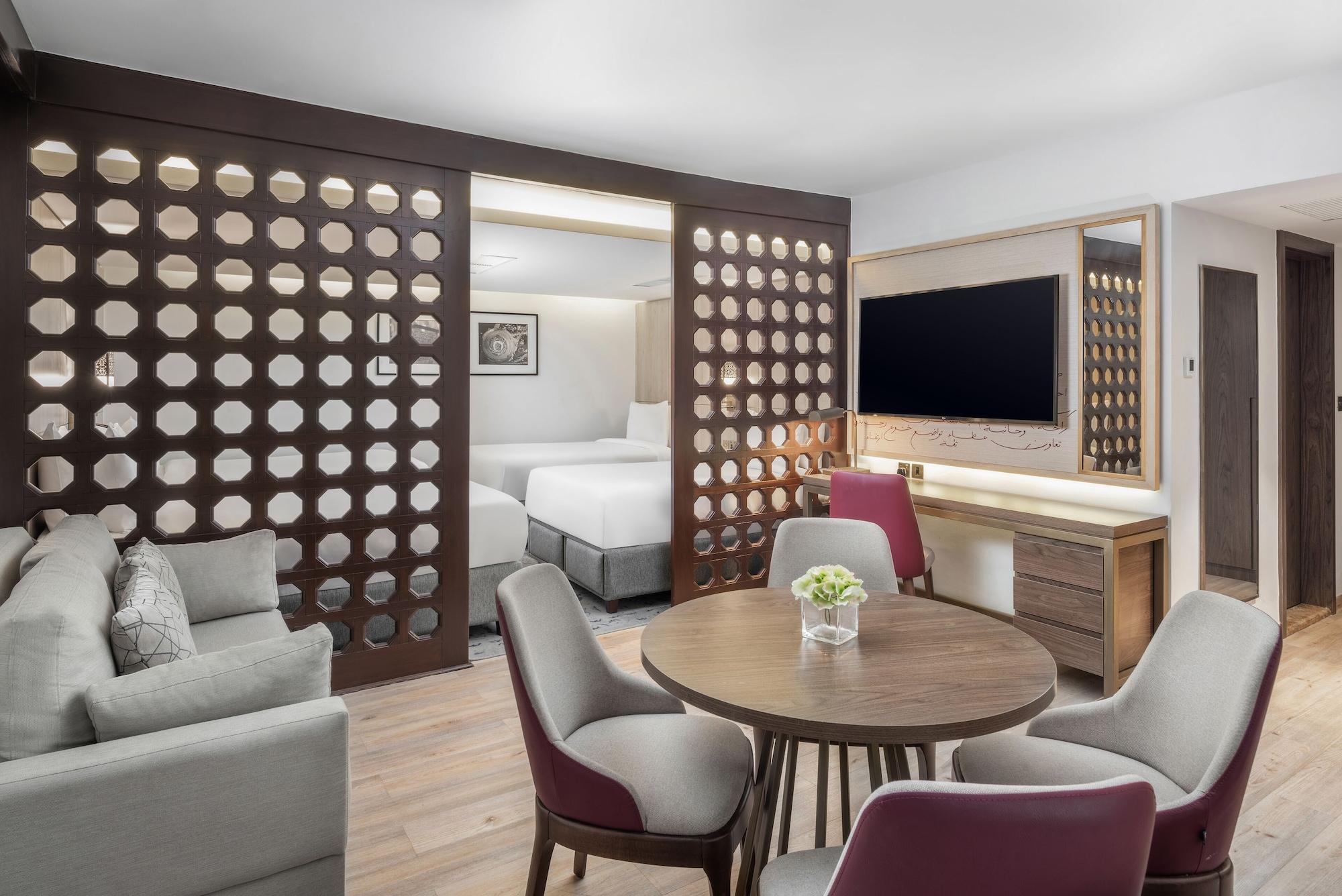 מכה Makarem Ajyad Makkah Hotel מראה חיצוני תמונה The photo shows a modern hotel room or suite. It features a living area with a comfortable gray sofa and a round wooden dining table surrounded by upholstered chairs. A large flat-screen TV is mounted on the wall opposite the seating area. There's a 