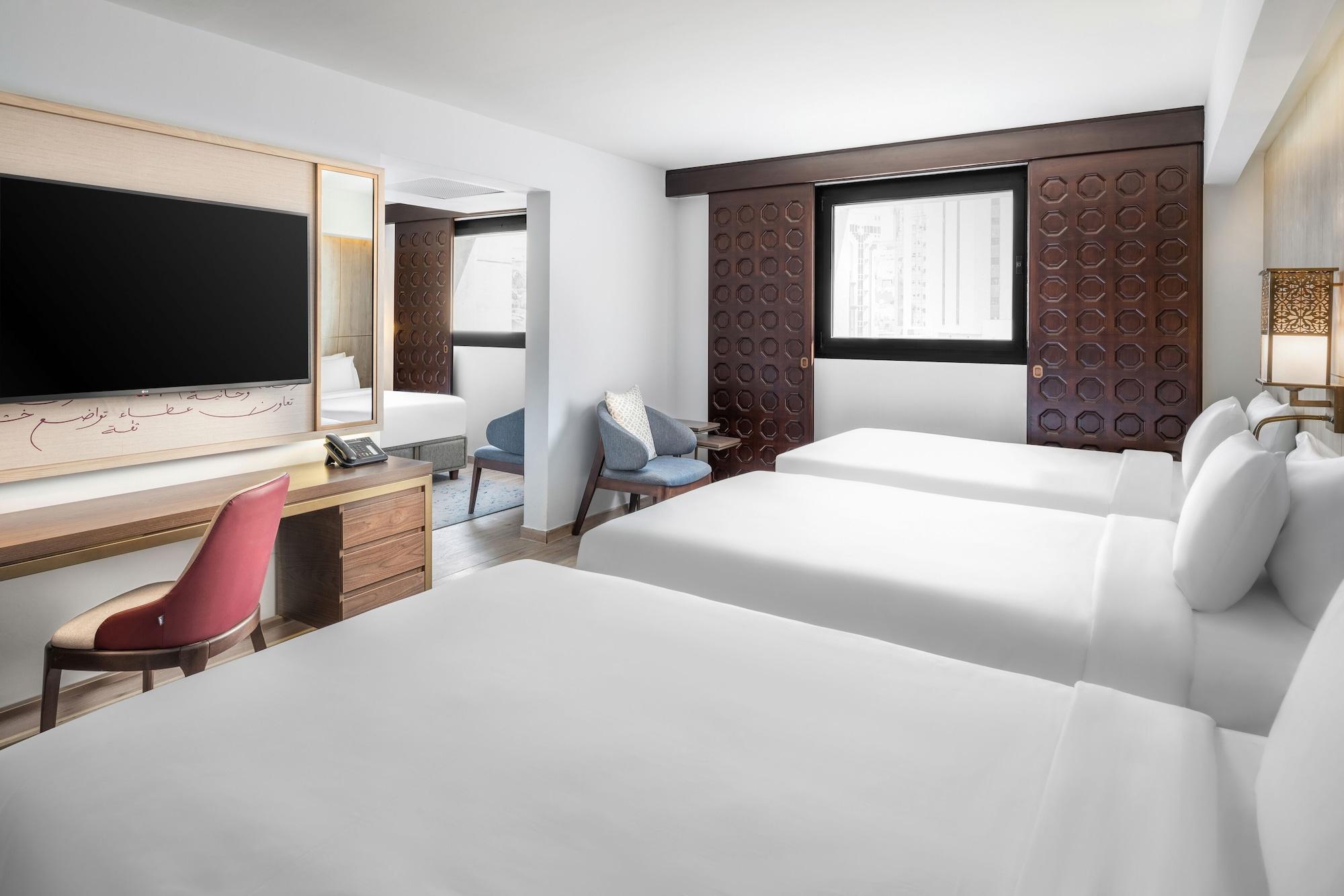 מכה Makarem Ajyad Makkah Hotel מראה חיצוני תמונה The photo shows a hotel room featuring three beds with white linens. There is a large flat-screen TV mounted on the wall opposite the beds, and a wooden side table beneath it. A red chair is positioned near the TV, while a blue upholstered chair is p