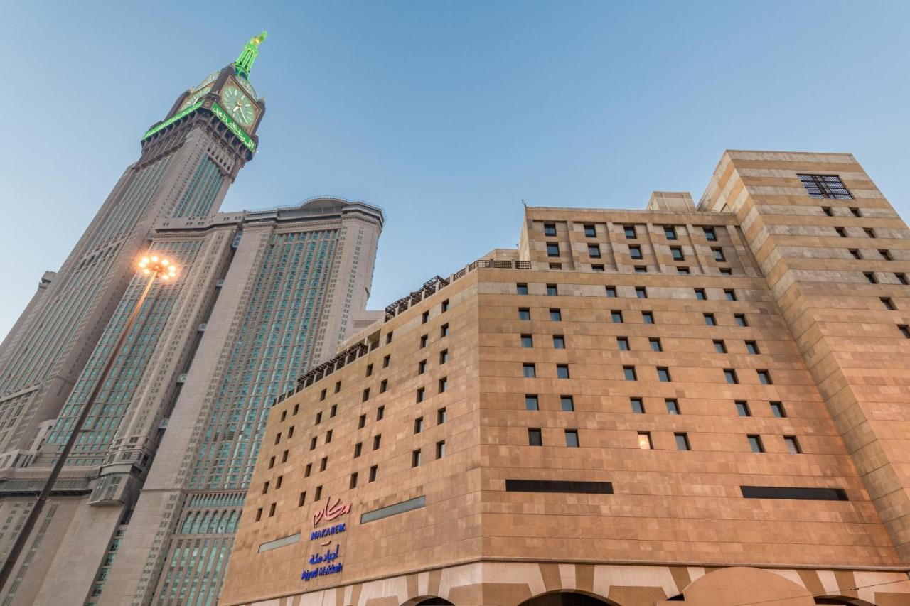מכה Makarem Ajyad Makkah Hotel מראה חיצוני תמונה The photo shows a prominent building with a modern architectural design, likely a hotel or commercial establishment, at the forefront. It features a light-colored stone facade with large windows. Behind it, there is a tall skyscraper, which has a dis