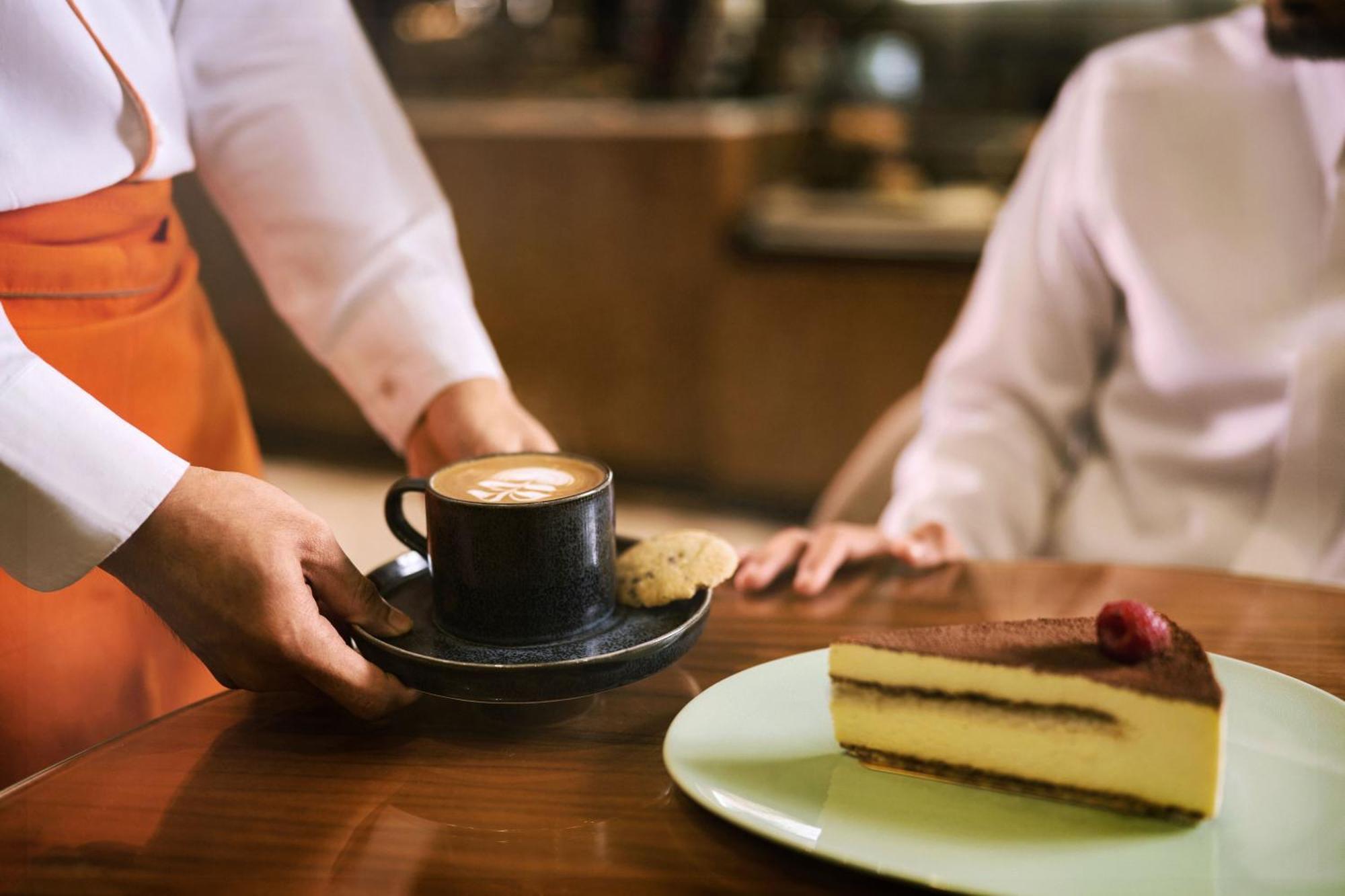 מכה Makarem Ajyad Makkah Hotel מראה חיצוני תמונה The photo shows a hand presenting a cup of coffee on a saucer, accompanied by a cookie. In the foreground, there is a slice of cake on a light green plate, garnished with a raspberry. The setting appears to be a café or restaurant, with a blurred bac