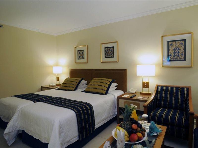 מכה Makarem Ajyad Makkah Hotel מראה חיצוני תמונה The photo shows a hotel room with a cozy and inviting atmosphere. It features two beds with white linens and dark striped bedspreads. There are two bedside lamps providing warm light, and the walls are decorated with framed artworks. Beside the beds 