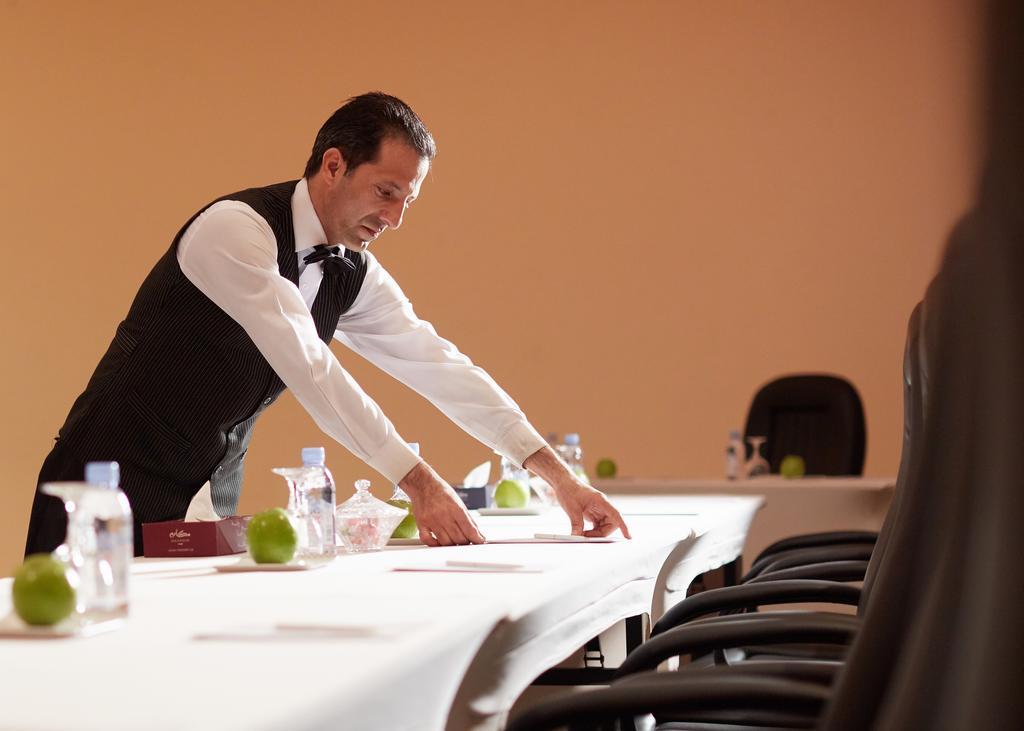 מכה Makarem Ajyad Makkah Hotel מראה חיצוני תמונה The image shows a waiter or service staff member in a formal attire, which includes a vest and bow tie. He is preparing a long table, likely for an event or meeting. The table is set with items such as water bottles and green apples, and there are se
