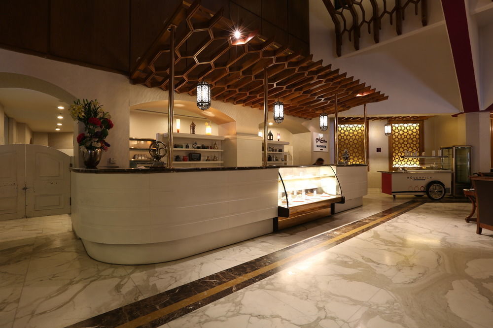 מכה Makarem Ajyad Makkah Hotel מראה חיצוני תמונה The photo shows a modern reception area or lobby of a hotel or restaurant. The focal point is a curved front desk made of white material, featuring a glass display case that showcases various items. Above the desk, there is a decorative wooden struct